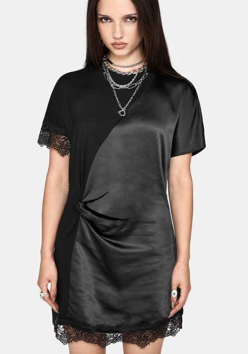 A woman modeling a black satin-like short-sleeve dress with lace trim at the sleeve cuffs and hem, layered with silver chain necklaces, against a light gray studio background.