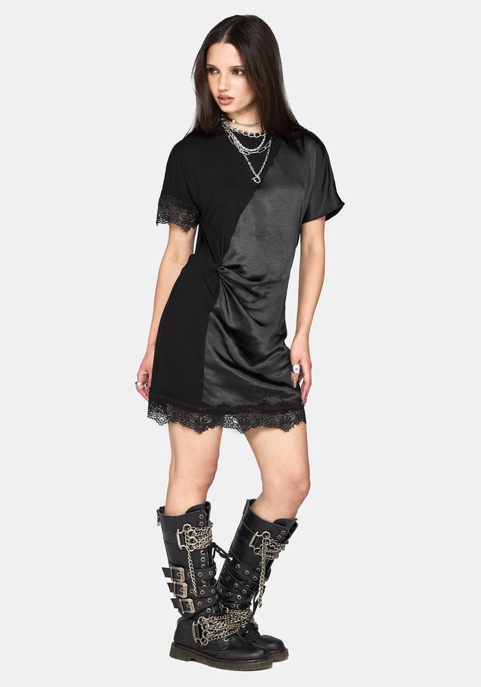 A model wearing a black short-sleeve dress with a satin-like sheen, lace trim, and a twisted detail at the waist, paired with layered silver necklaces and black lace-up combat boots, against a light gray background.