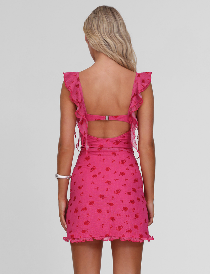 A woman with long blonde hair is shown from the back wearing the TIGERMIST AMARIS MINI DRESS - DITSY FLORAL, which features a pink V-neck, red floral patterns, ruffled sleeves, and an open back with a horizontal strap. She wears a white bracelet.
