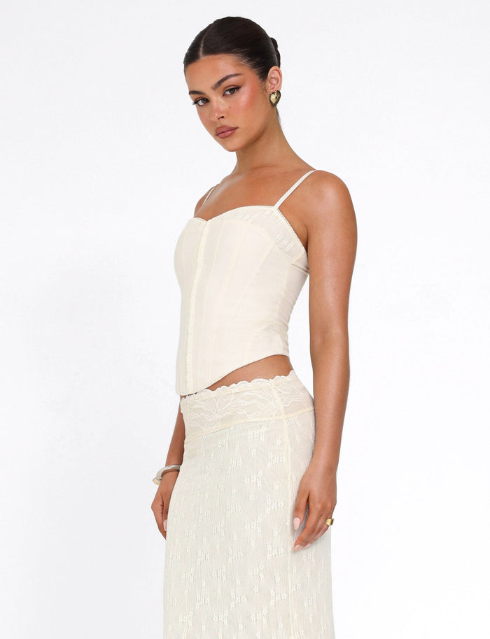 A woman wears the TIGERMIST ANTONELLA TOP in cream, featuring delicate lace panels, paired with a matching lace skirt. She stands against a plain white background, looking slightly to the side with a neutral expression.