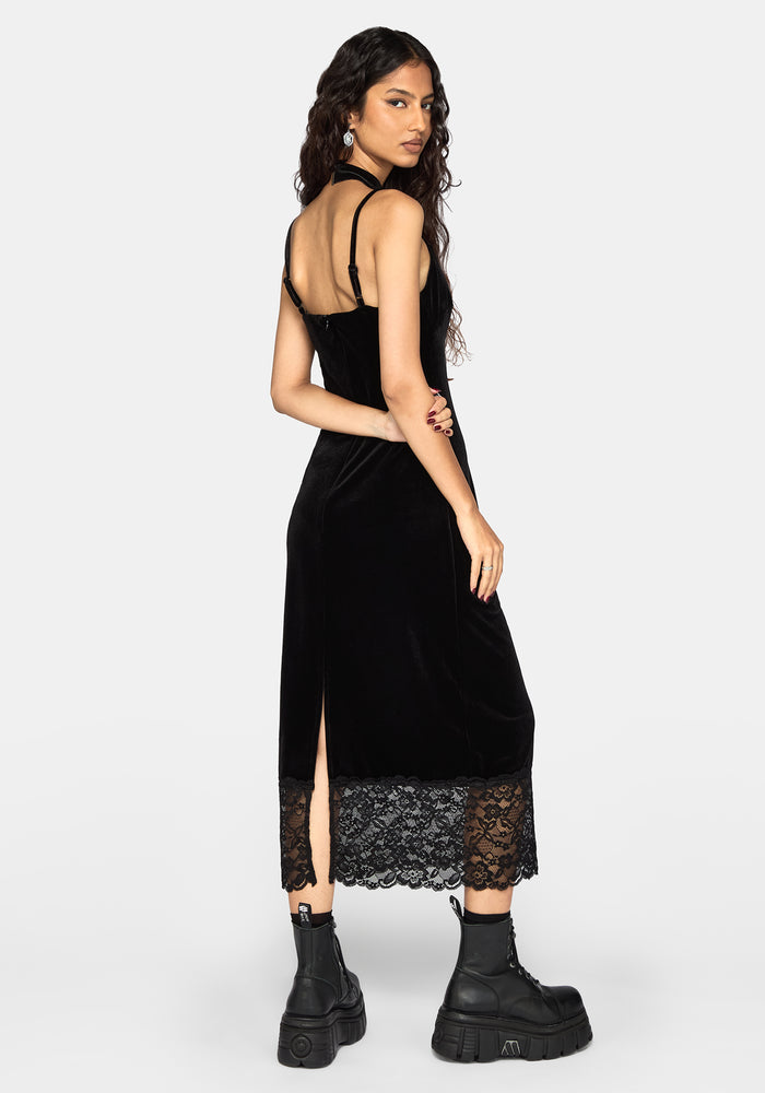 A woman modeling a black velvet midaxi dress with thin straps, a v-neckline, and a lace hem, shown from the back against a light gray studio background.