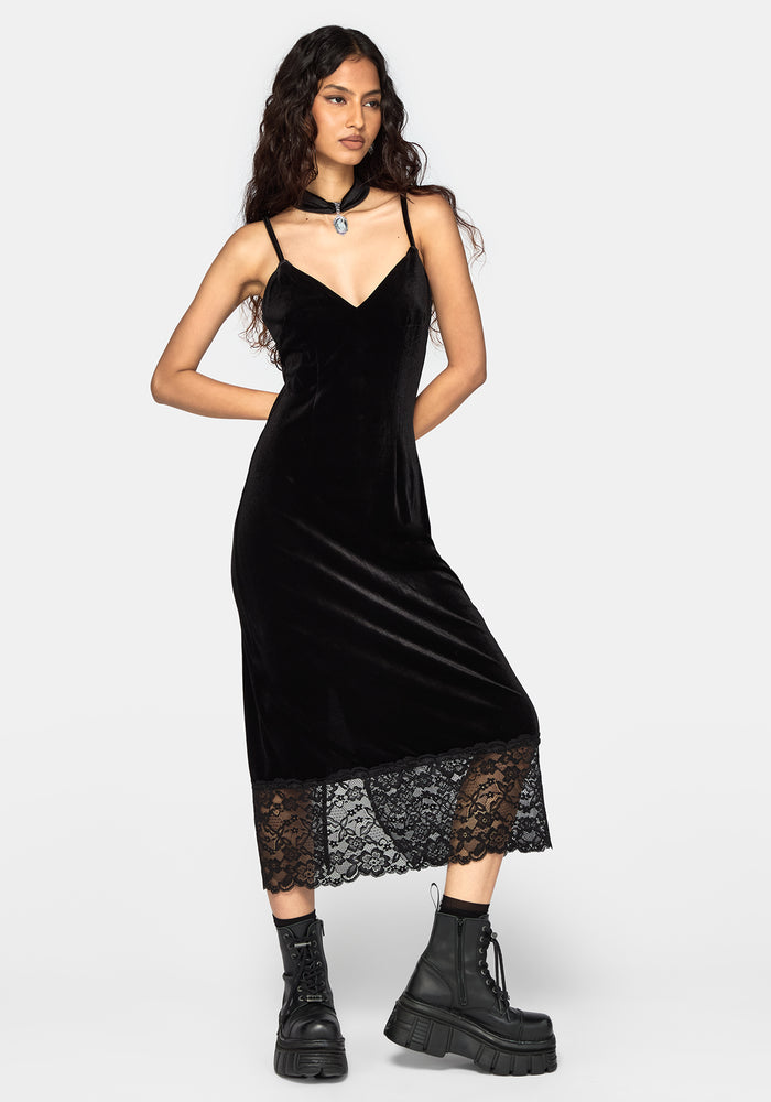 A woman modeling a black velvet midi dress with thin straps, a sweetheart neckline, and a black lace hem, styled with a choker necklace and black combat boots against a light gray background.