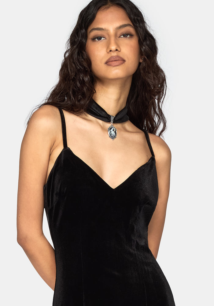 A woman modeling a black velvet slip dress with thin straps and a matching black choker necklace featuring a silver-toned pendant, set against a plain light gray background.
