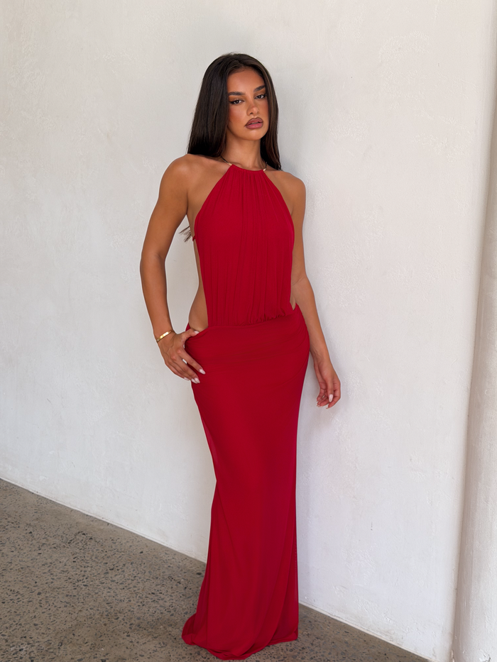 A woman with long dark hair models the TIGERMIST EVANNA MAXI DRESS - RED, a sleeveless, floor-length high neck dress, standing against a white textured wall and gazing confidently at the camera.