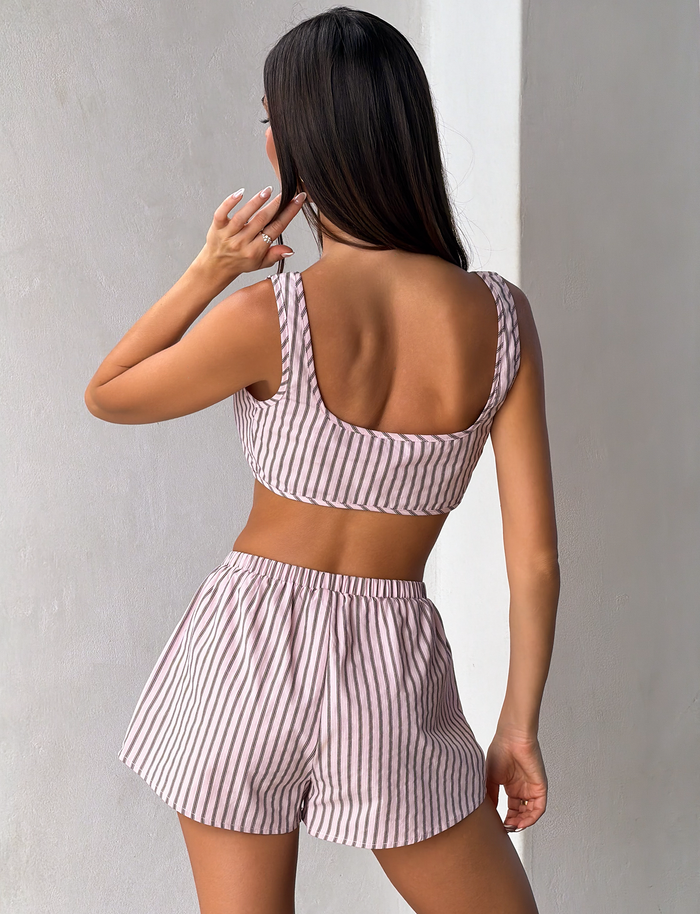 A woman with long brown hair stands against a light wall, facing away with one hand near her head, wearing the TIGERMIST EZRA TOP - PINK STRIPE and matching shorts.