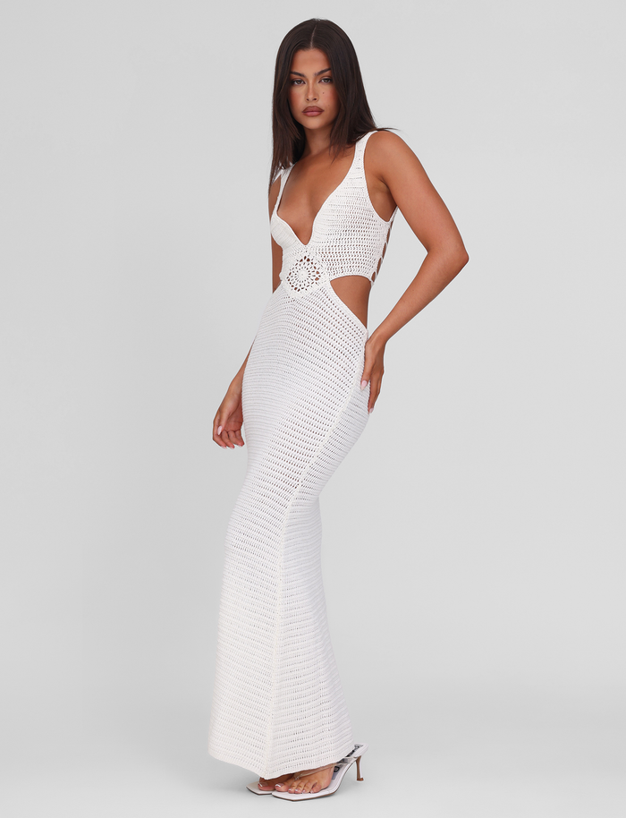 A woman models the TIGERMIST JONAH MAXI DRESS in cream—a fitted, sleeveless crochet dress with waist cutouts, deep V-neckline, and flared hem—paired with white heeled sandals against a plain light background.