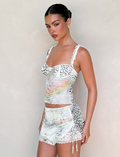A woman stands against a light wall wearing the TIGERMIST JOSEPHINE MINI SKIRT in white animal print with matching lace-trimmed camisole, her hair styled in a sleek bun and accessorized with pearl earrings.