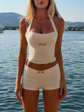 A woman wears the TIGERMIST KITTIE BUCKLE TOP in cream and matching shorts as she stands by the water, with boats and mountains visible in the background on a sunny day.