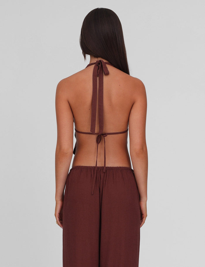 A person with long brown hair, seen from the back, wears the TIGERMIST LAINEY TOP - CHOCOLATE linen halter tied at neck and back, paired with matching loose brown pants against a plain light gray background.