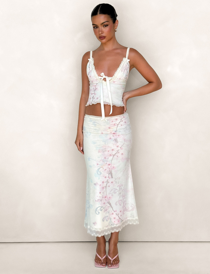 A woman with dark hair styled back and a neutral expression wears the TIGERMIST LONIE MIDI SKIRT in Neutral Floral Washed Cherry Blossom, paired with a pastel floral sleeveless top and pink heeled sandals against a plain light background.