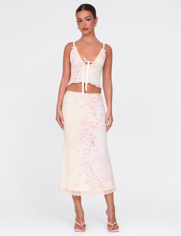 A woman wears the TIGERMIST LONIE MIDI SKIRT in Washed Cherry Blossom floral print with a matching pale, sleeveless tie-front top, styled simply with open-toed heels and minimal jewelry against a plain white background.