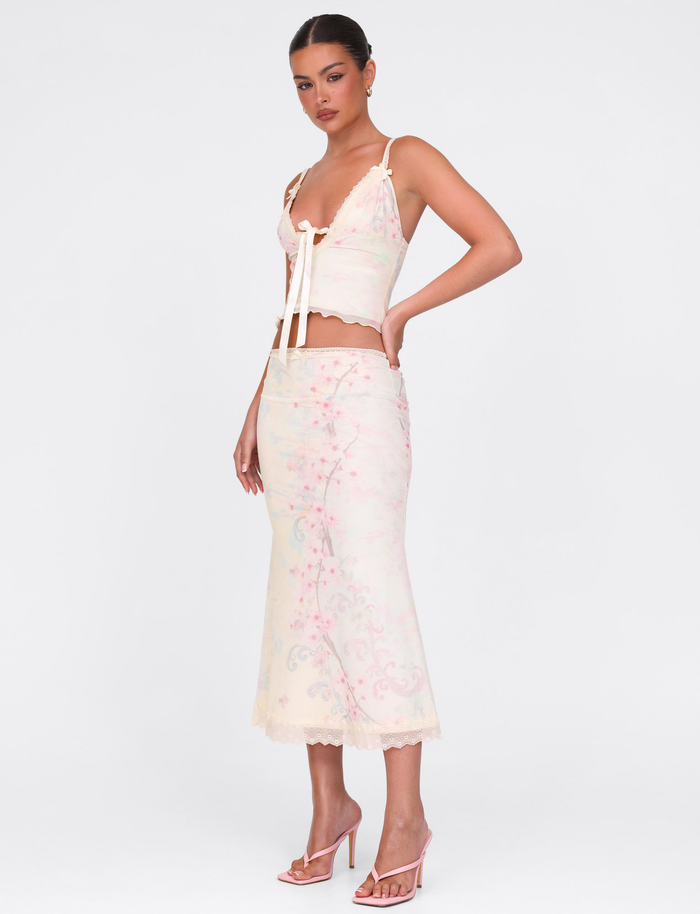 A woman in profile wears the TIGERMIST LONIE MIDI SKIRT - NEUTRAL: FLORAL: WASHED CHERRY BLOSSOM, a pastel floral mesh midi skirt with an exclusive print and matching sleeveless lace-trimmed top. Her hair is tied back and she has strappy high-heeled sandals.