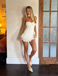 A woman wears the TIGERMIST LYRA MINI DRESS in cream, featuring a corset bodice and ruffle details, paired with matching cream heels. She stands indoors by a wooden door with frosted glass, her head slightly tilted and one leg bent, while soft lamp light illuminates the room in the background.
