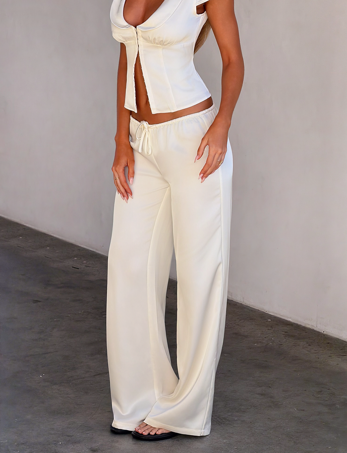 A woman stands against a plain wall with her hands by her sides, wearing the TIGERMIST MAURA PANT in white. She pairs the loose, wide-legged pants with a sleeveless white crop top featuring tie details. Her face is not visible in the image.