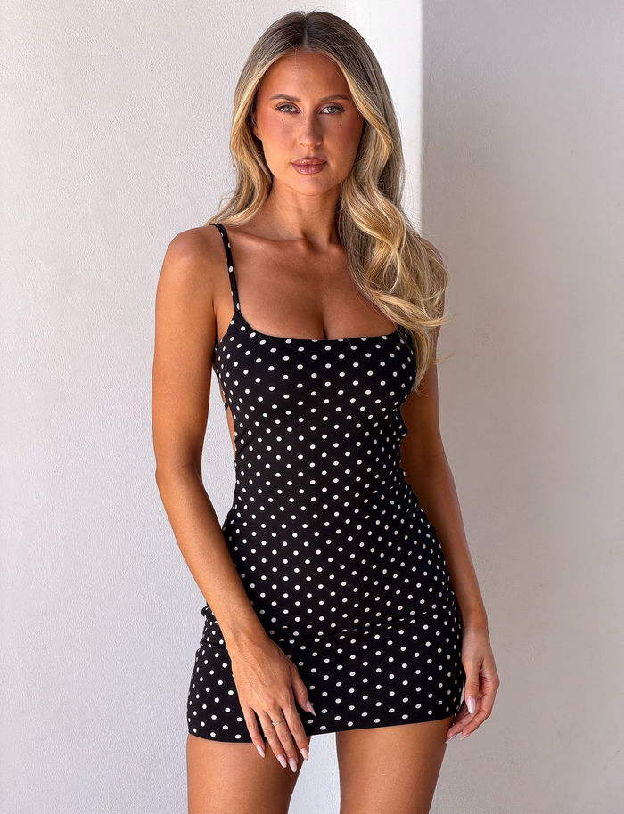 A woman with long blonde hair models the TIGERMIST MINERVA MINI DRESS 2.0 in black polka dot, featuring adjustable shoulder straps, as she stands against a light-colored wall and looks at the camera with a neutral expression.
