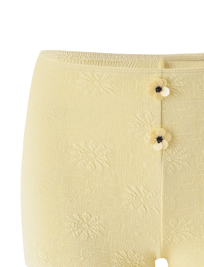 Close-up of TIGERMIST's QUINLAN SHORT - LEMON: light yellow mini shorts with a subtle floral pattern, delicate floral trim, and two small flower-shaped buttons with black centers on the front waistband.