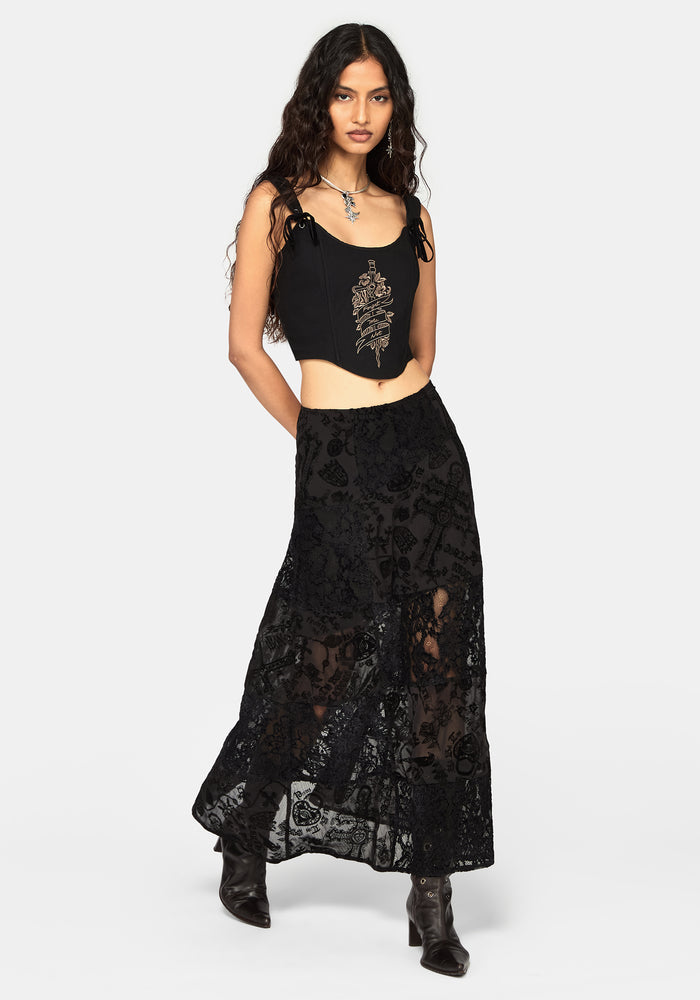 A model wearing a black sleeveless corset top with gold embroidery and a matching black lace maxi skirt, paired with black ankle boots, against a light gray studio background.