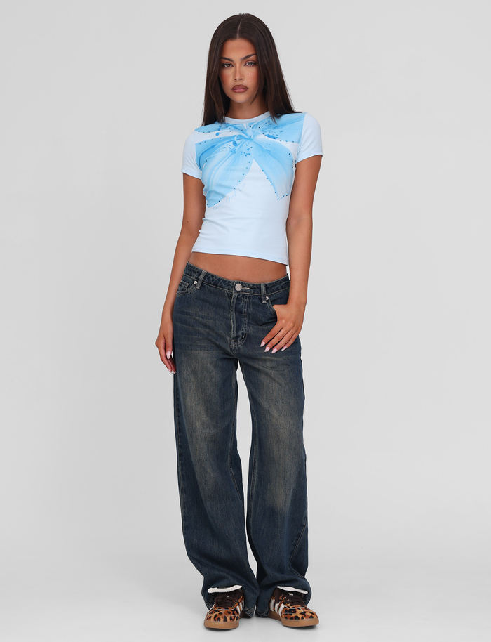 A woman with straight brown hair wears the TIGERMIST SAPHIRA TEE in blue, featuring a butterfly design and stretchy fabric, paired with baggy dark blue jeans and leopard print shoes, standing against a plain white background.