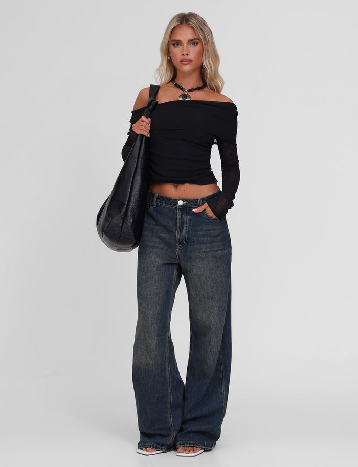 A woman stands against a plain background wearing the TIGERMIST VERA TOP in black, loose blue jeans, black sandals, a black necklace, and carrying a large black bag over her shoulder.